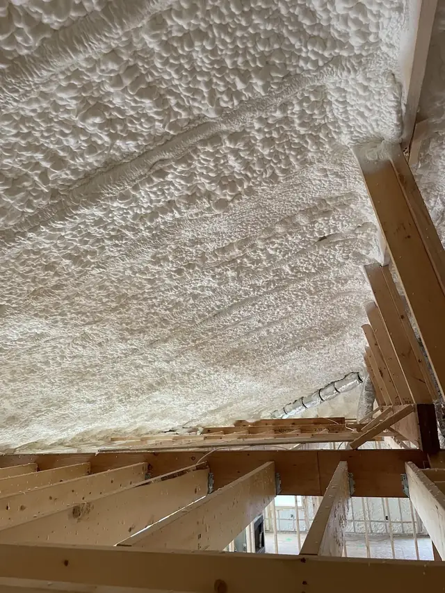 Underside of a roof deck coated in thick white spray foam, seen from the attic floor joists.