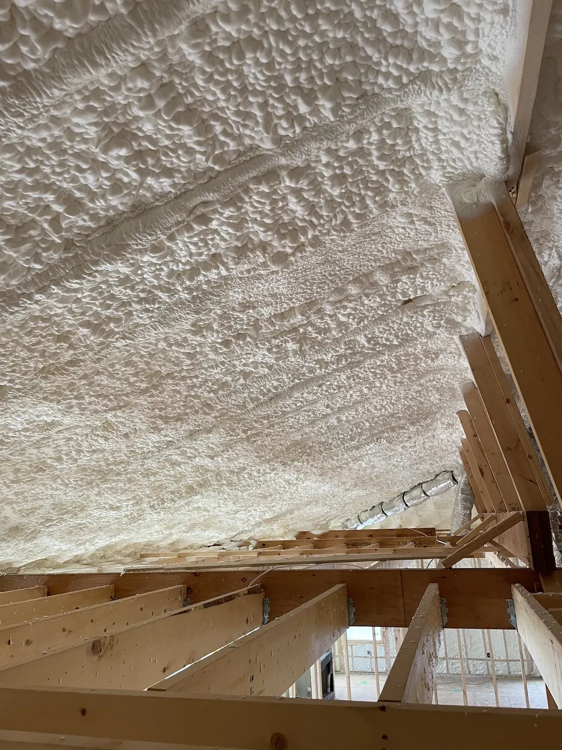 Underside of a roof deck coated in thick white spray foam, seen from the attic joists.