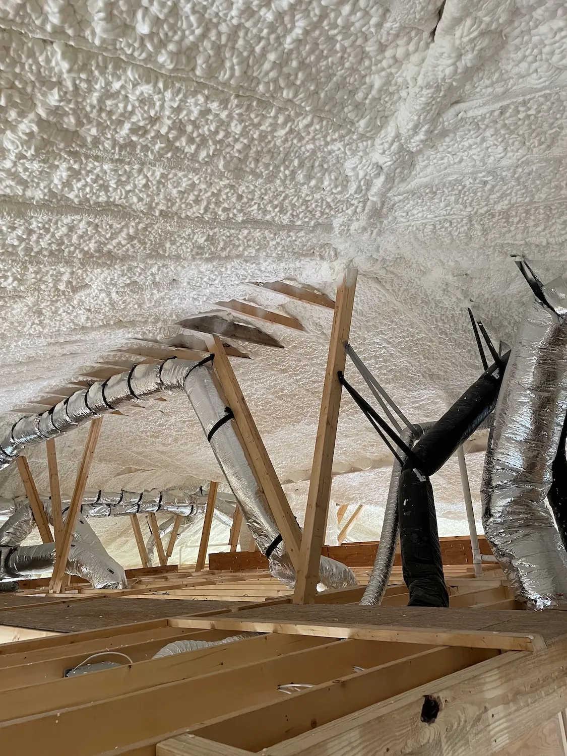 Attic roof deck coated in thick white spray foam, with insulated ductwork running between the rafters.