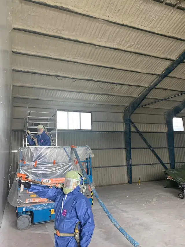 Technician on a scissor lift spraying foam across the ceiling panels of a steel commercial building.
