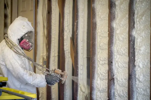 Technician in a hooded respirator suit spraying foam between the wall studs of a building under construction.