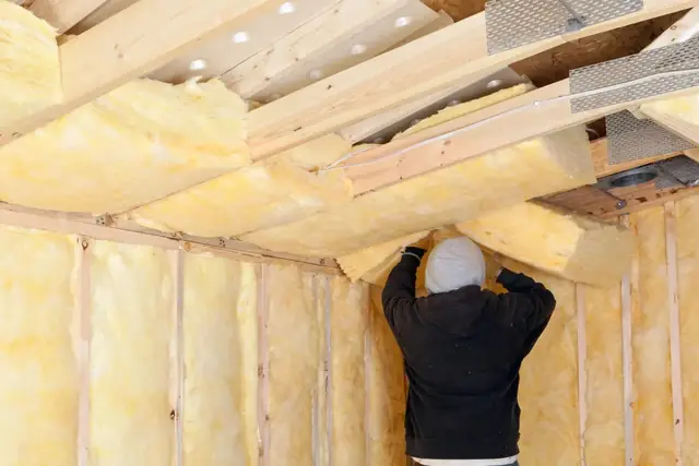 Worker fitting yellow fiberglass batt insulation between the ceiling joists of a room.