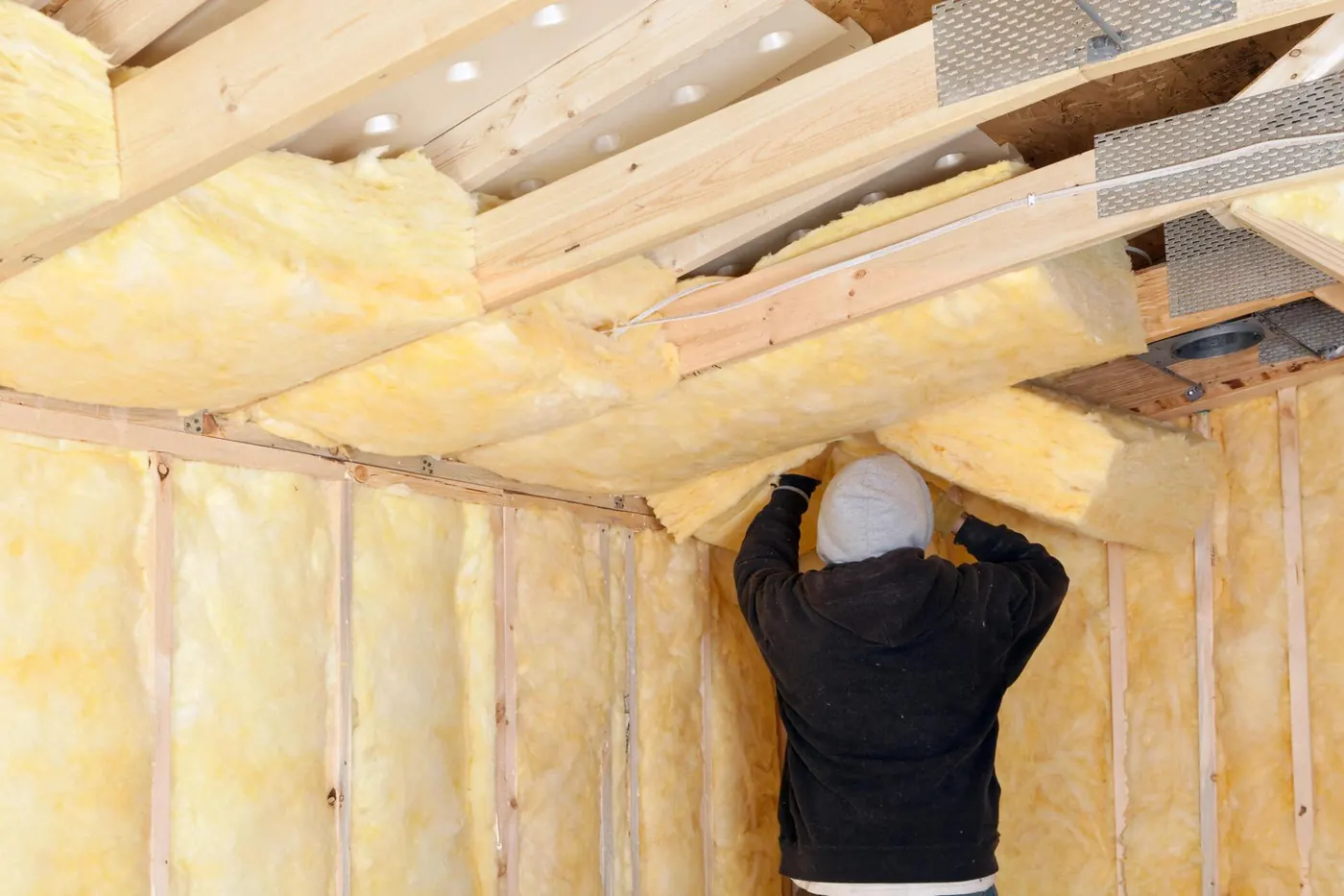 Worker fitting yellow fiberglass batt insulation between the ceiling joists of a room.