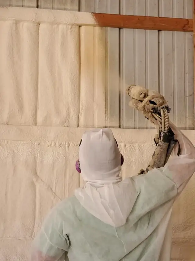 Technician in protective gear spraying foam onto the interior wall panels of an agricultural building.