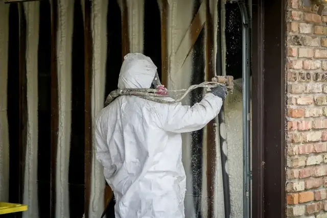 Technician in a full protective suit spraying foam into a wall cavity beside a brick exterior wall.