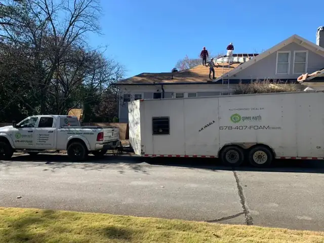Crew working on a house roof beside the Green Earth spray foam trailer parked in the driveway.