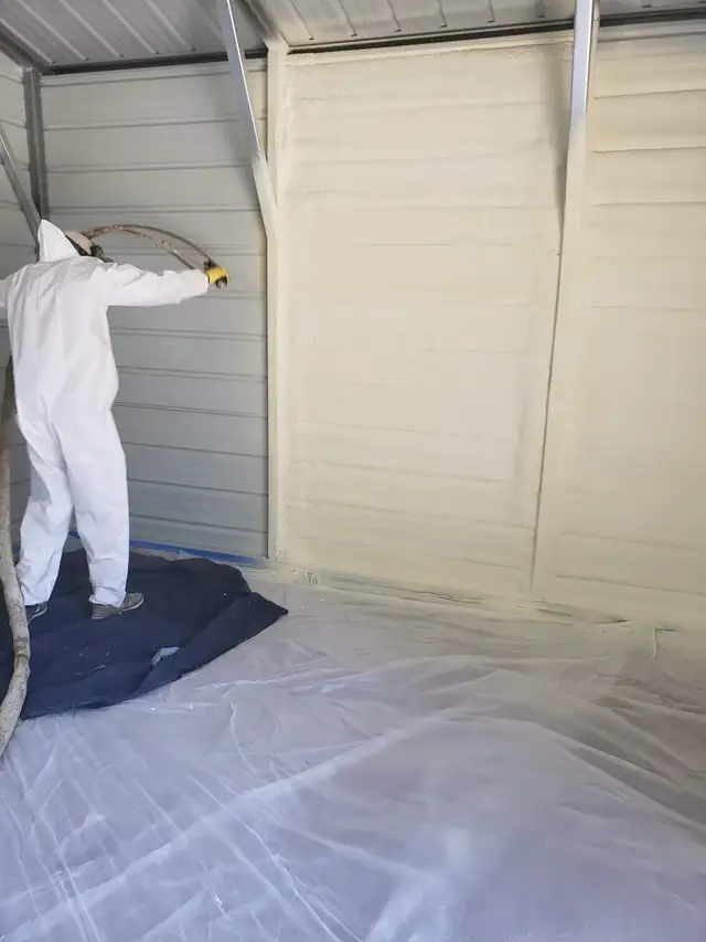 Technician spraying foam onto the interior wall of a steel building, with sheeting laid over the floor.