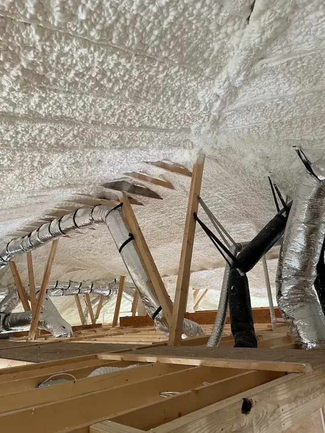 Attic roofline fully coated with white spray foam, with flexible ductwork running between the rafters.